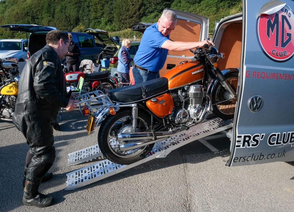 Two men load a classic orange Honda motorcycle into a silver camper van RV