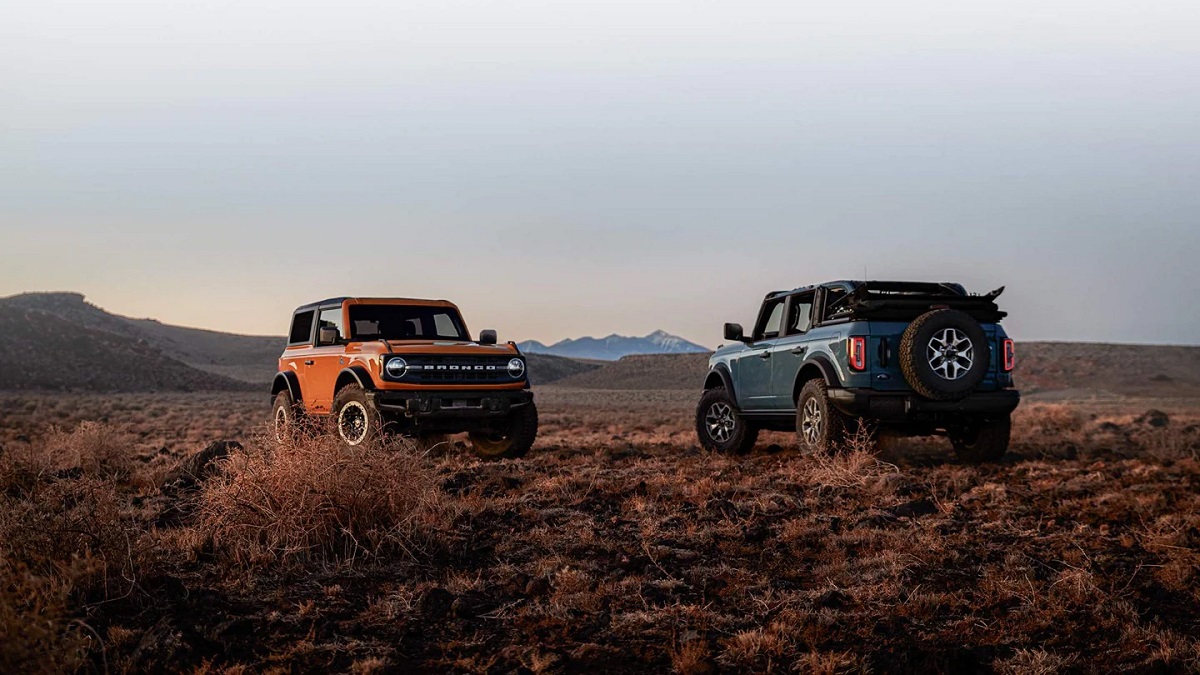 An orange and a blue Ford Bronco sit on a plain with mountains in the background.