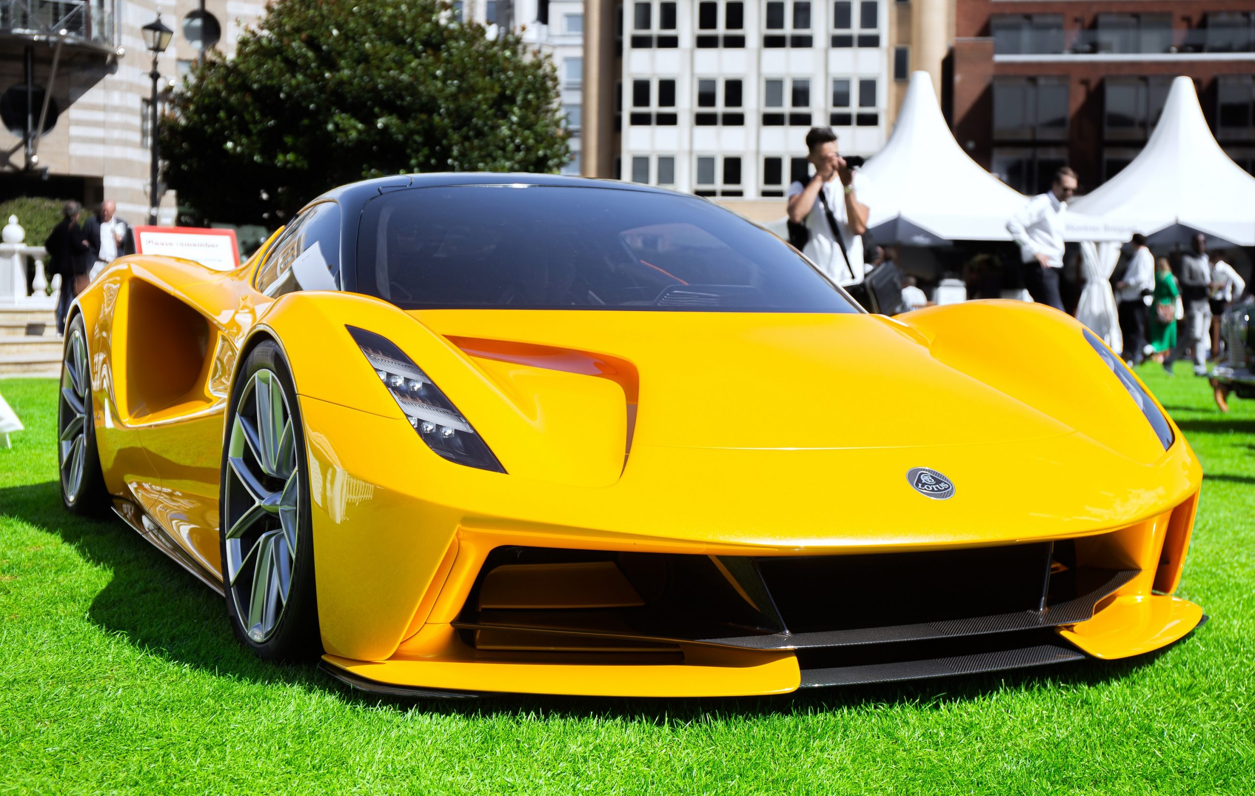 A yellow Lotus Evija, the brand's electric hypercar, shot from the front 3/4 on a concourse lawn