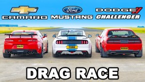 A red chevrolet camaro (left) next to a ford mustang (middle) and a dodge challenger (right) all preparing for a drag race.