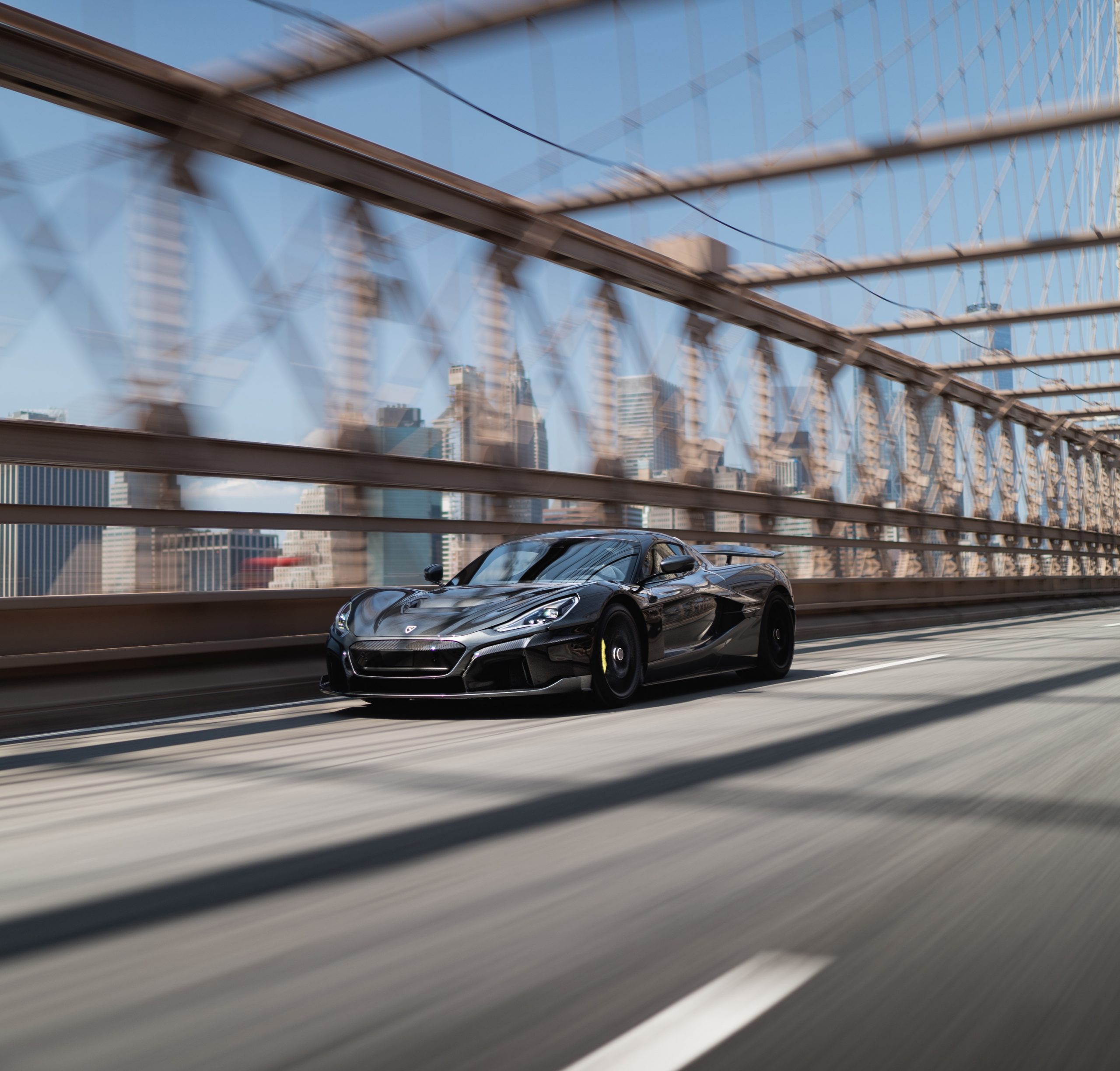 A grey Rimac Nevera, arguably the king of the electric supercars, drives down a bridge shot from the front 3/4.