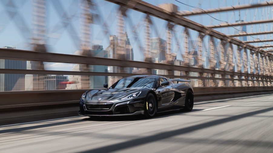 A grey Rimac Nevera, arguably the king of the electric supercars, drives down a bridge shot from the front 3/4.