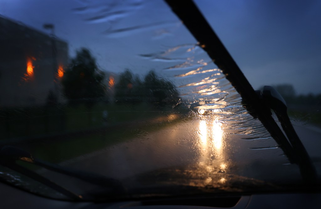 Behind a windshield wiper, a car drives along a road in heavy rain. 