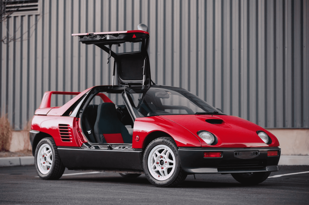 A red Autozam AZ-1 from 1992 is parked with the right gullwing door open