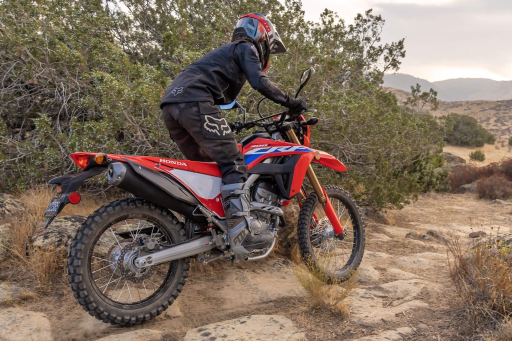 The rear 3/4 view of a black-clad rider on a white-red-and-blue 2021 Honda CRF300L riding through the desert