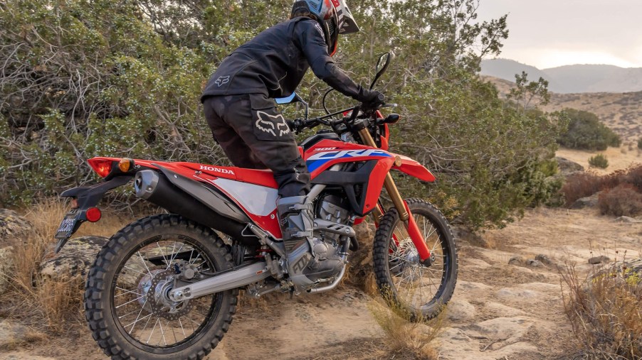 The rear 3/4 view of a black-clad rider on a white-red-and-blue 2021 Honda CRF300L riding through the desert