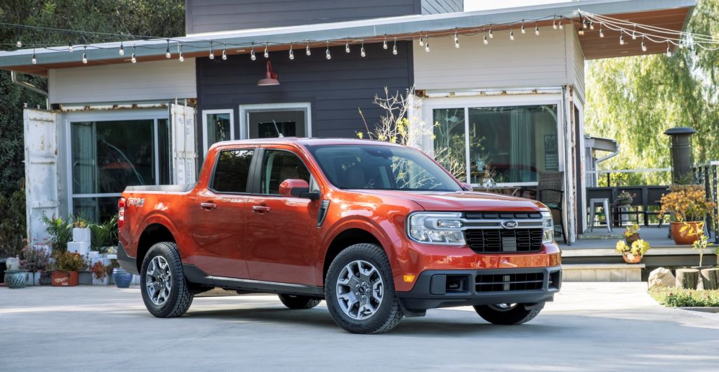 The 2022 Ford Maverick Lariat compact pickup truck in orange red parked outside of a house and patio, it either has the hybrid or all-gas EcoBoost engine.