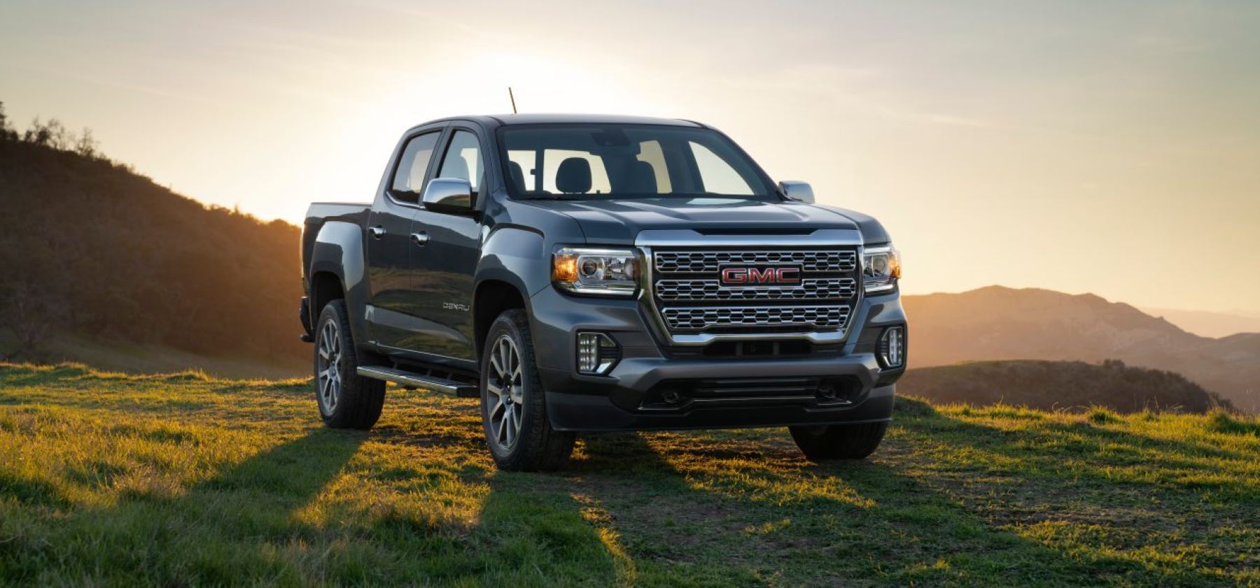 The 2022 GMC Canyon midsize pickup truck parked on a grass field as the sun sets on rolling hills