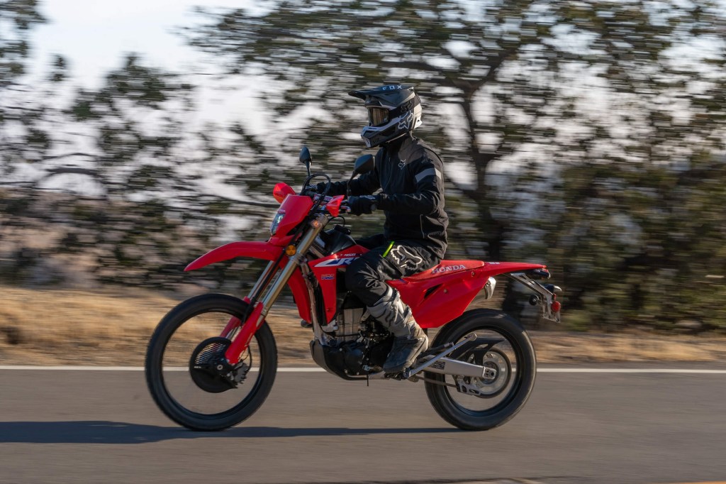 A black-clad rider takes a red 2022 Honda CRF450L dirt bike down a paved road