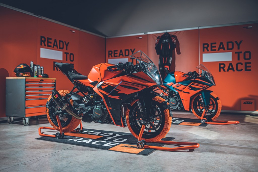Two orange-and-black 2022 KTM RC 390s with tire warmers on paddock stands in a garage