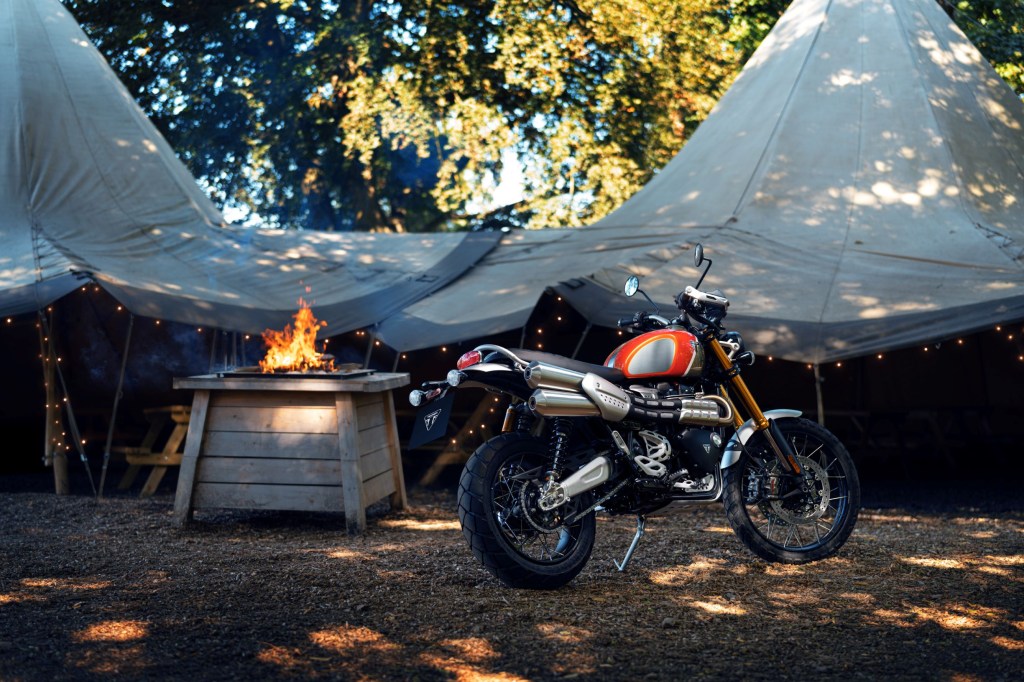 The rear 3/4 view of an orange-silver-and-gold 2022 Triumph Scrambler 1200 XE Gold Line in the forest amongst some canvas tents