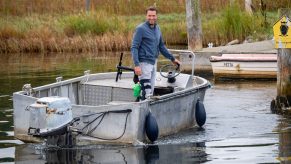 A white boat approaching a dock near a grassy area driven by a man with a blue shirt and white pants.