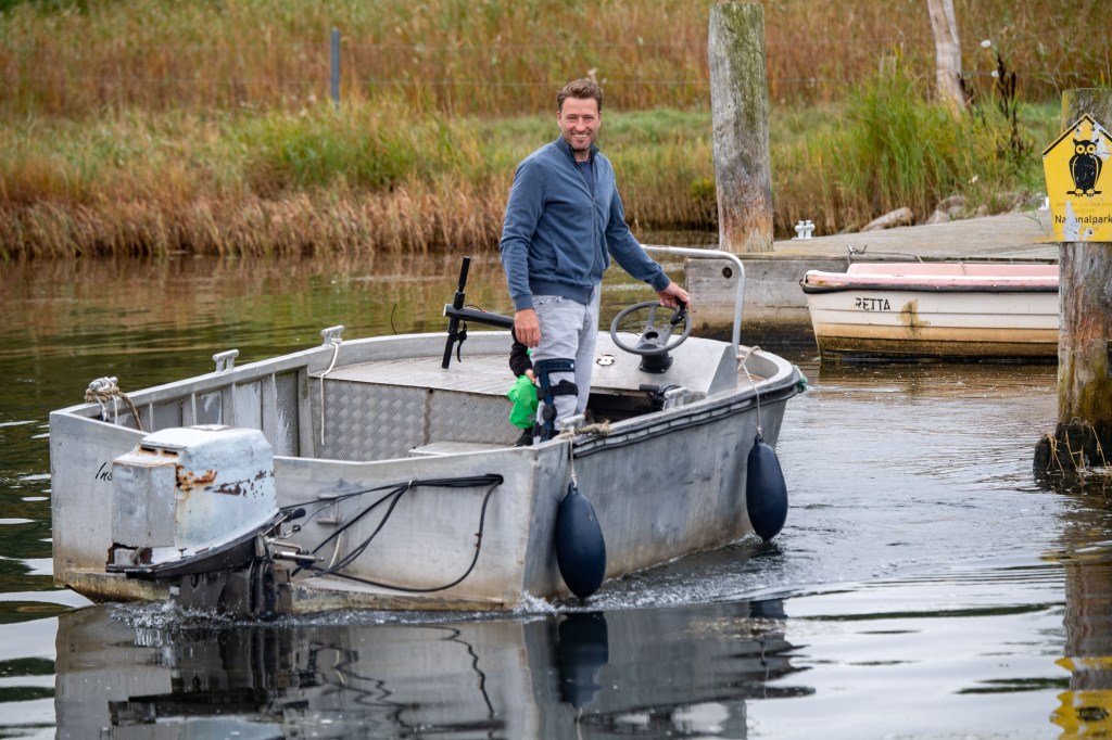 A white boat approaching a dock near a grassy area driven by a man with a blue shirt and white pants.