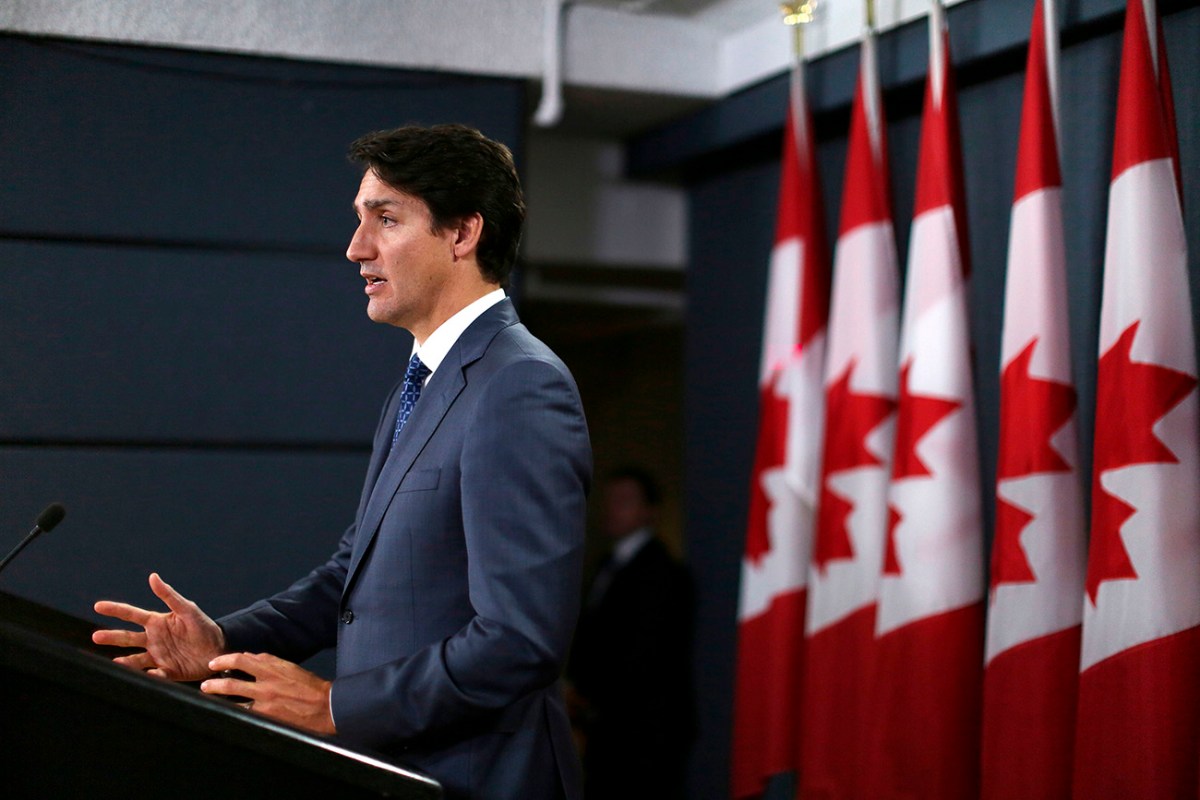 Canadian Prime Minister Justin Trudeau speaking at a podium facing toward the left of the frame with a row of five Canadian flags behind him