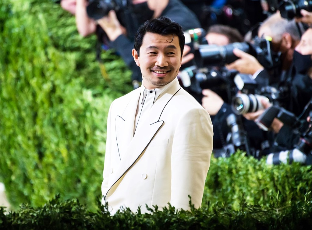 Simu Liu in a white suite with photographers in the background surrounded by shrubbery. 