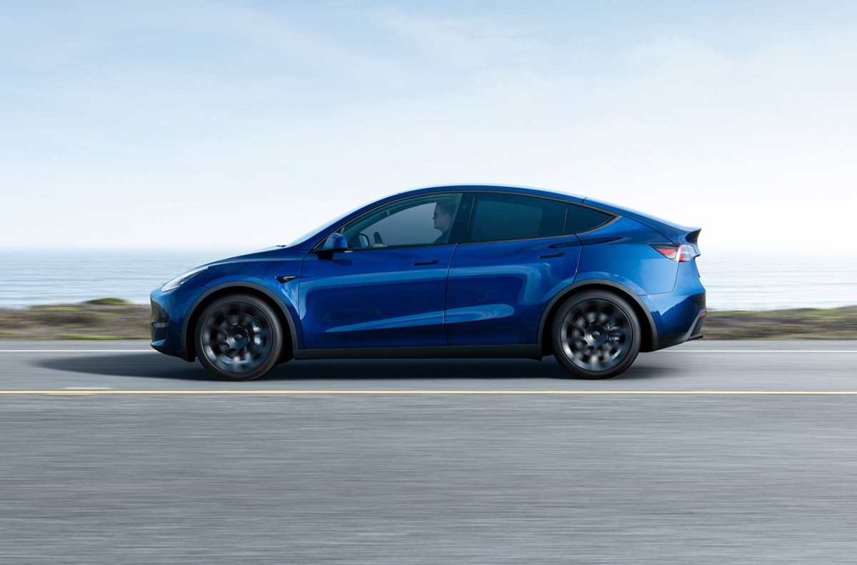 Tesla Model Y in blue shot from the side as it drives long a road. The Model Y is part of a recent Tesla suspension recall