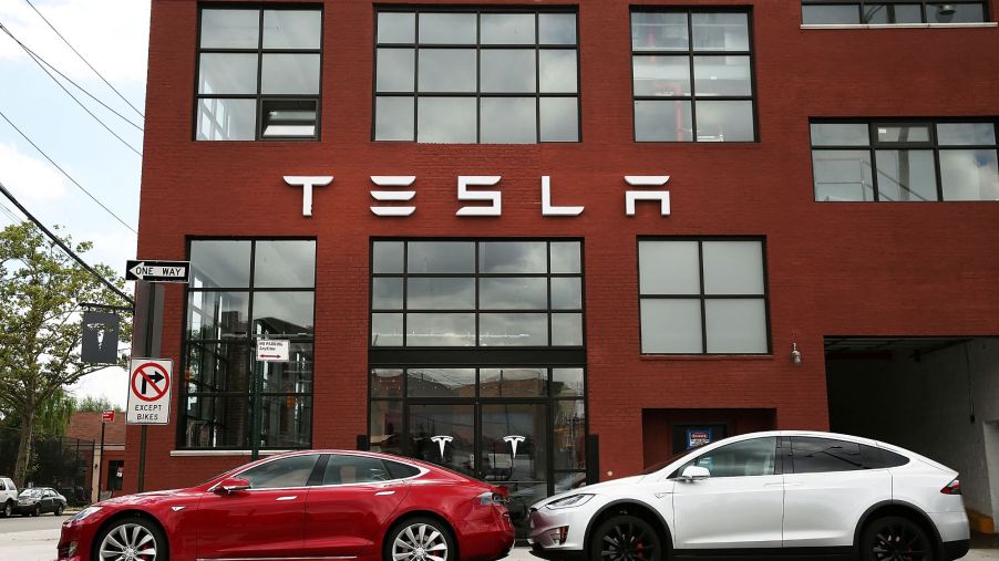 A red building with Tesla written on it with two Tesla cars, those capable of autopilot, in front of the building.