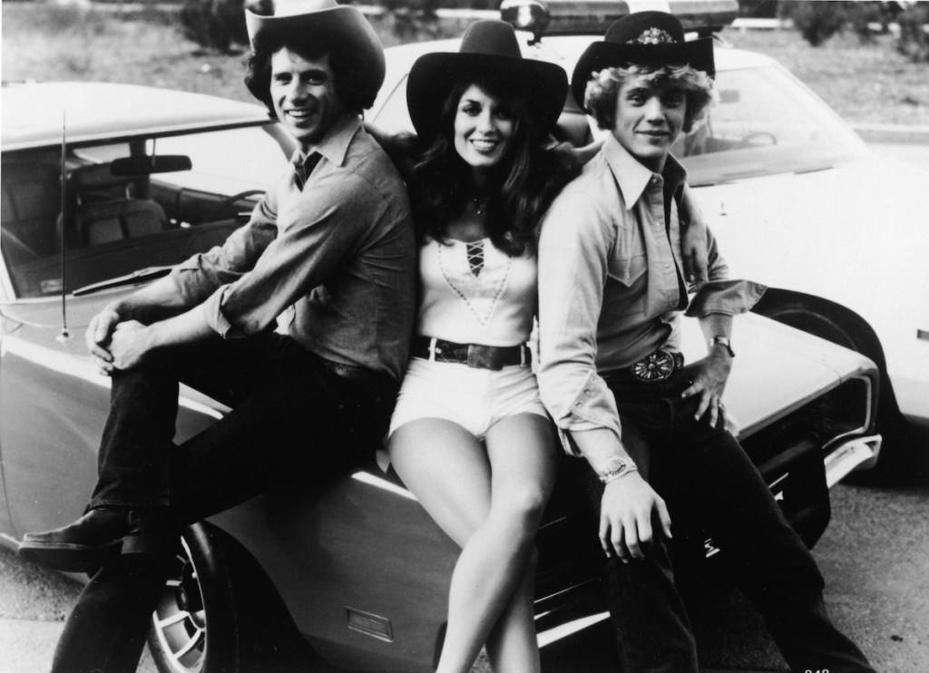 'The Dukes of Hazzard' stars Tom Wopat (left), Catherine Bach, and John Schneider sit on the hood of the General Lee circa 1980