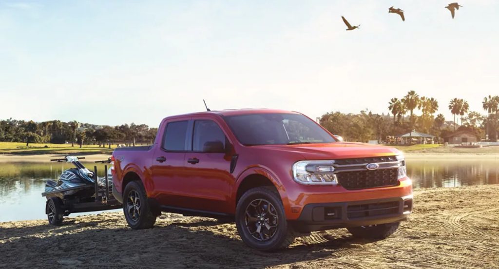 A red 2022 Ford Maverick compact pickup truck is towing two jetskis near a lake. 