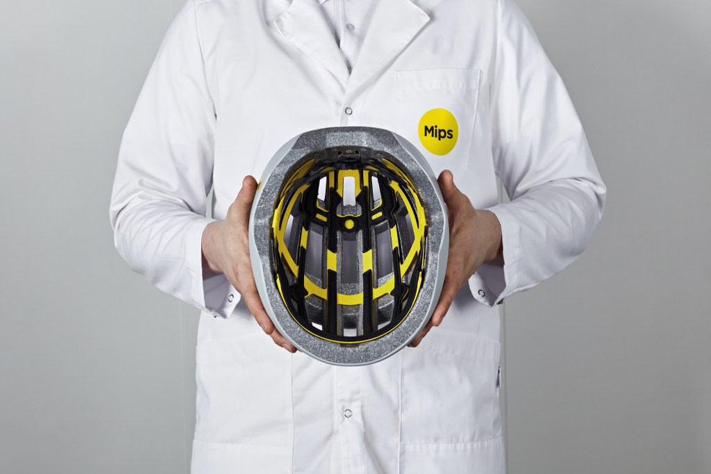 An individual in a white lab coat holds a gray bike helmet with a black-and-yellow MIPS liner