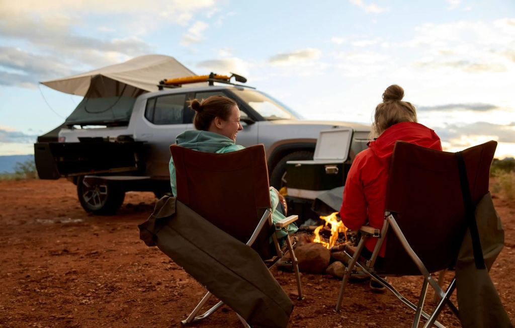 Two woman relaxing by a fire near a silver Rivian R1T