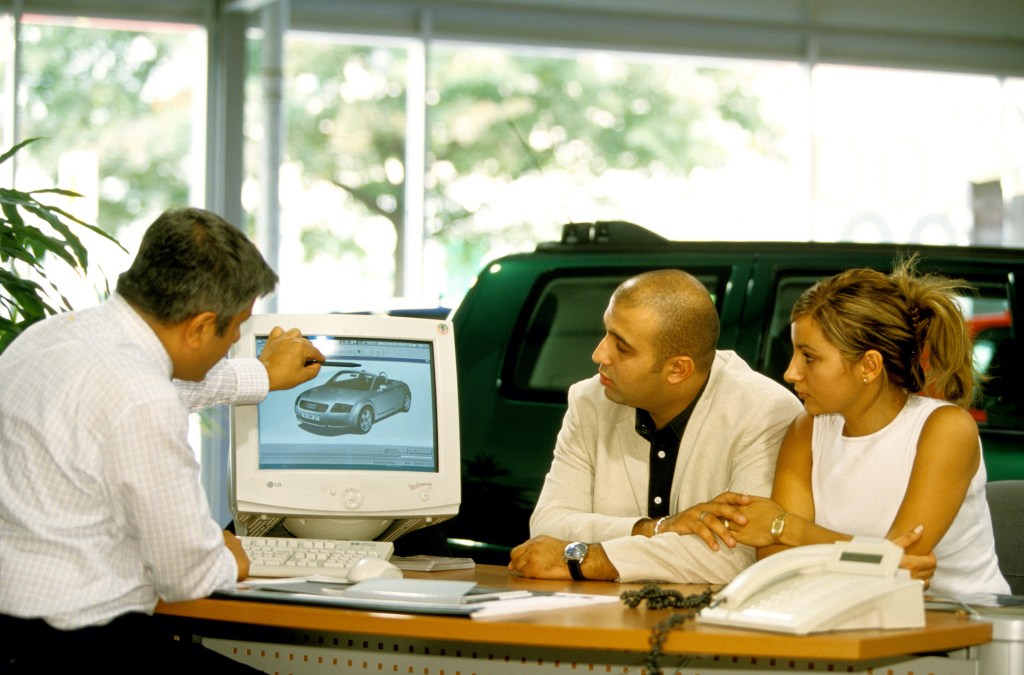  A car salesperson tells a couple of customers about the TT while sitting at his desk.
