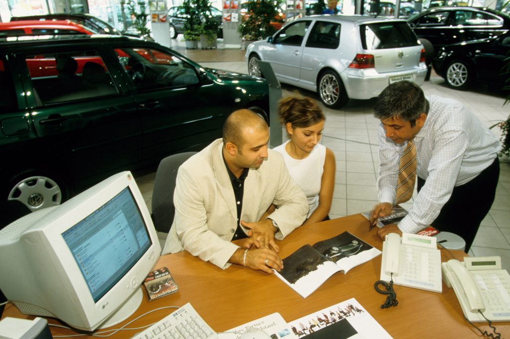 A car salesperson talks to a couple of customers about the Audi TT at his desk.