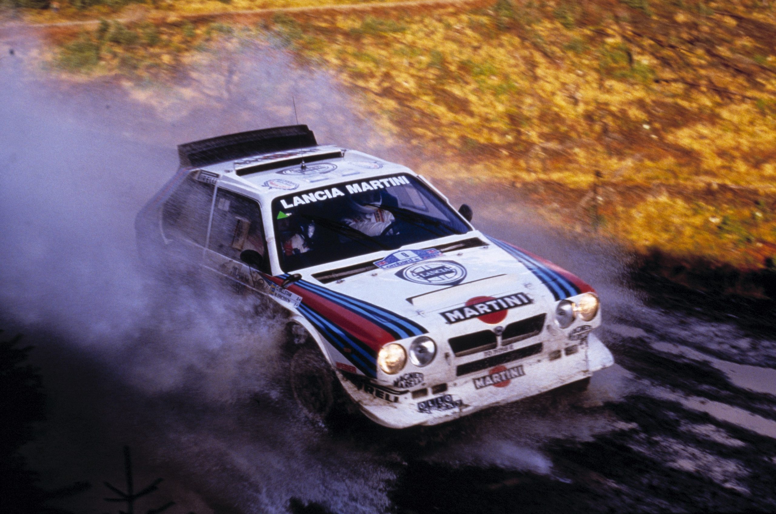 A blue and white Martini livery Lancia Delta S4 rally car racing through a muddy puddle on a rally stage.