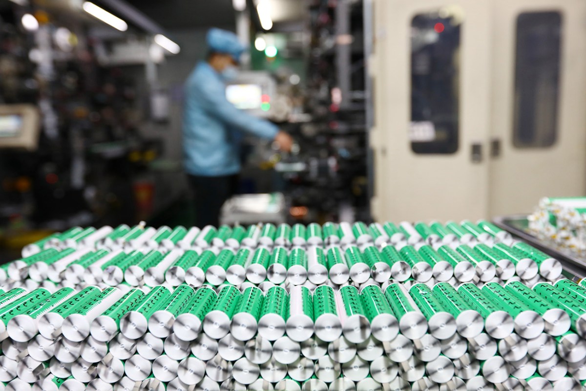 A stack of lithium ion batteries with green wrappings, in the blurred background an employee wearing a blue uniform and hat is working on assembling more battery cells