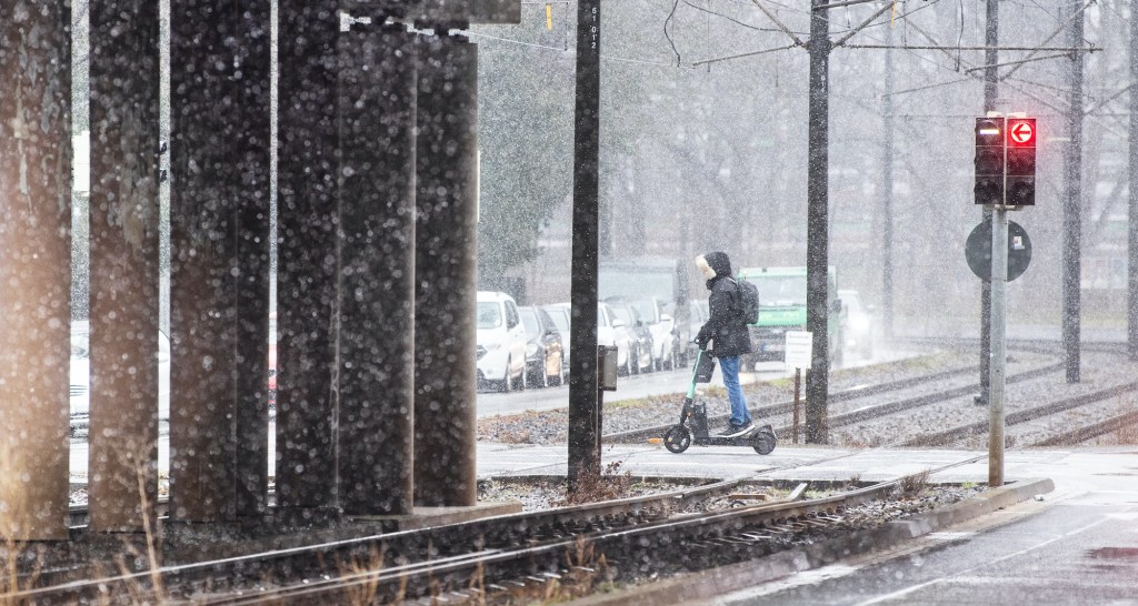 A man rides an e-scooter across light rail tracks in snowy conditions.