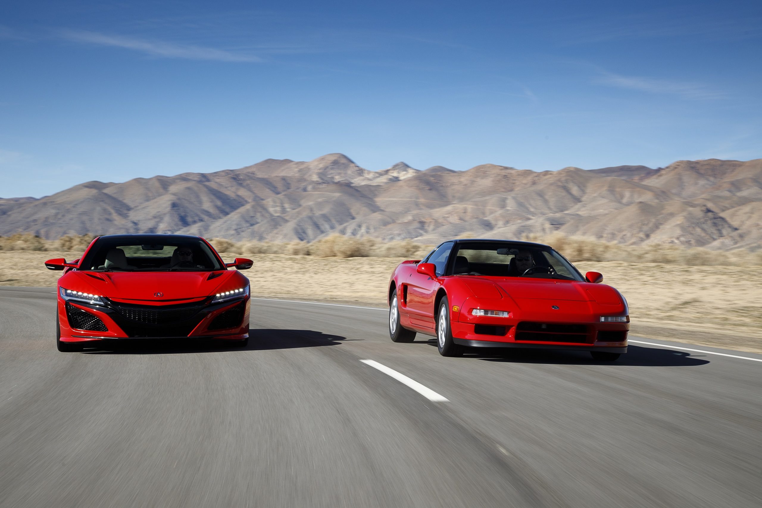A pair of red Acura NSX models, one new and one old, shot from the front while in motion