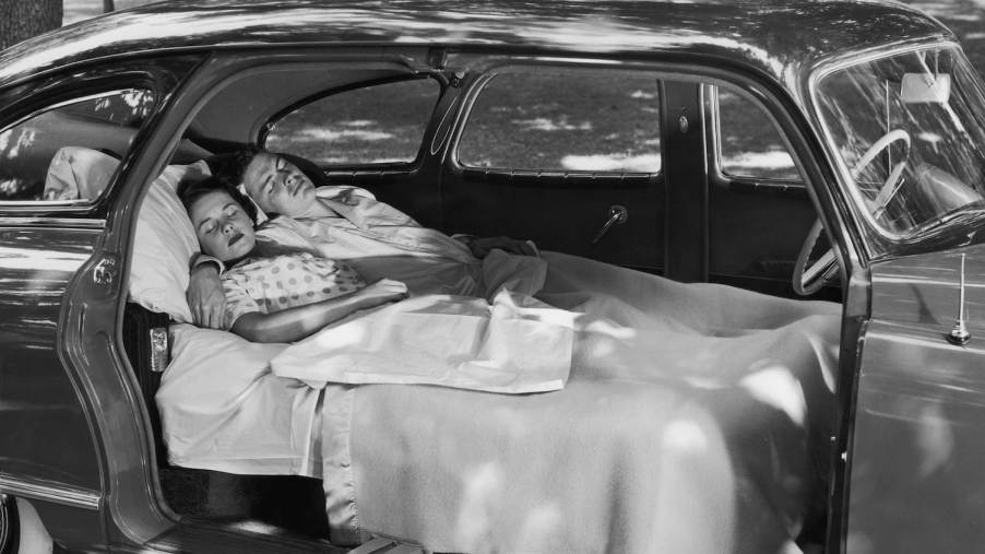 A couple sleeps in an antique car with fold-flat seats in a residential neighborhood.