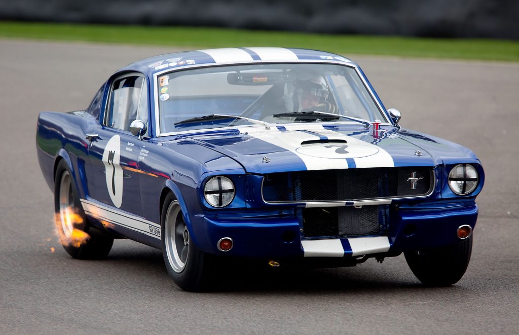 A blue-with-white-stripes 1965 Ford Mustang Shelby GT350 spitting flames while racing at the 2017 Goodwood Member's Meeting