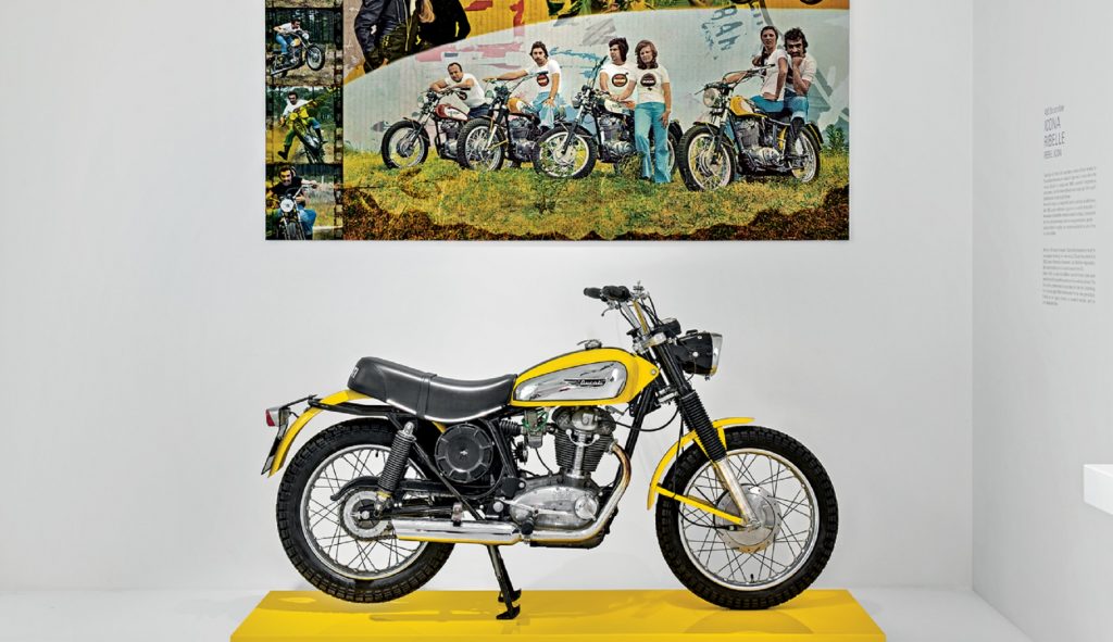 The side view of a yellow-and-silver 1969 Ducati 450 Scrambler in the company's museum in front of a photo of vintage Scrambler riders