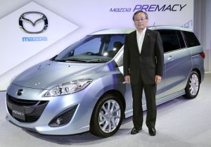 A silever 2010 Mazda 5 and Takashi Yamanouchi standing in front of it with a white background and the Mazda logo. 
