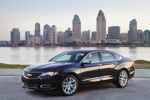 The 2018 Chevrolet Impala midsize sedan in black parked in front of a river and a city skyline of skyscrapers