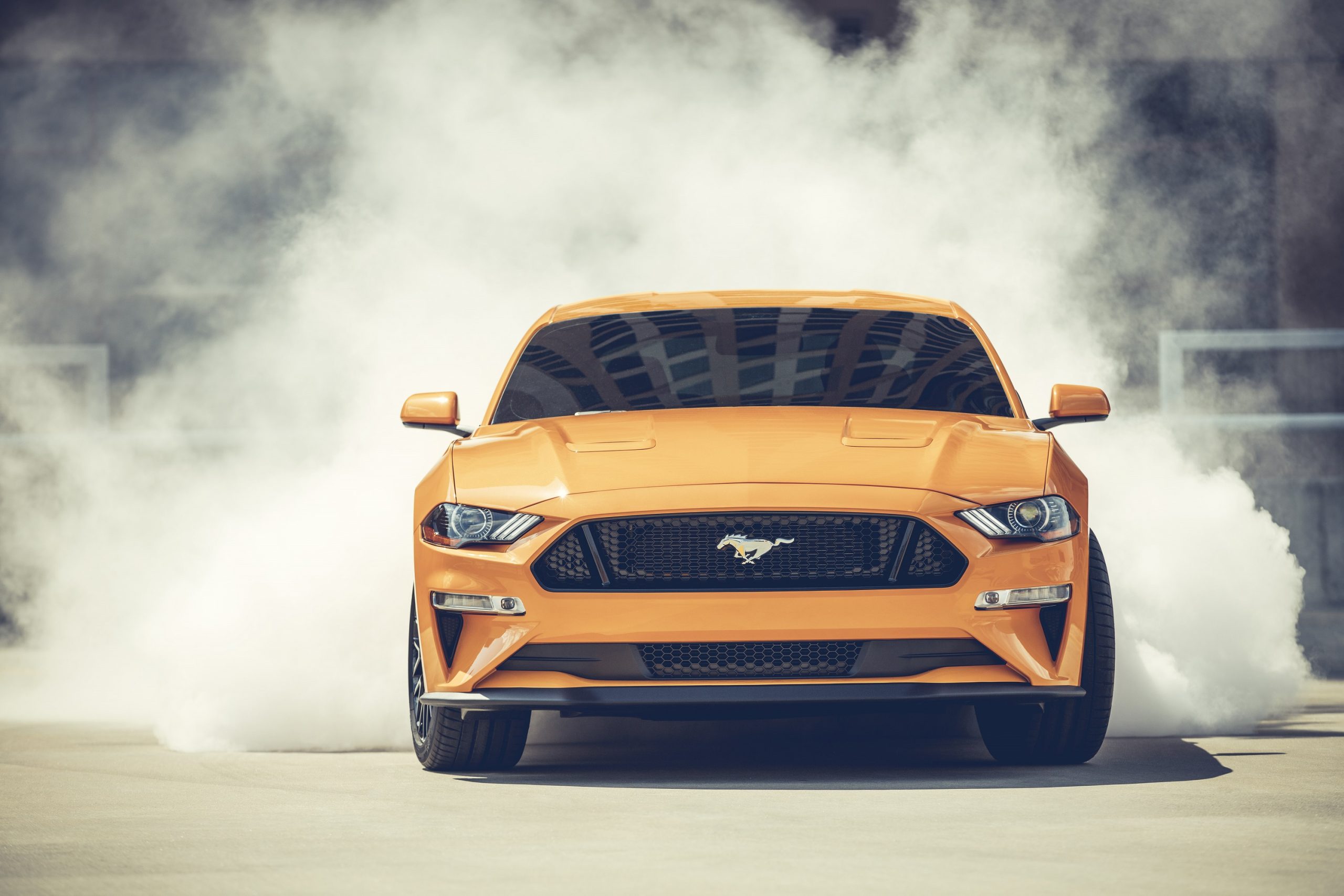 An orange Ford Mustang sports car mid-burnout shot from the front