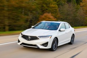 The 2021 Acura ILX A-Spec subcompact luxury executive car with a white paint color option driving on a stretch of country highway past a forest