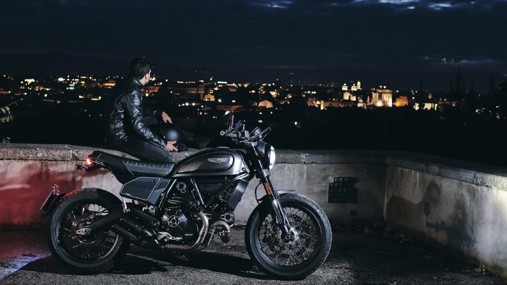 The side view of a black-clad rider sitting next to a black-and-gray 2021 Ducati Scrambler Nightshift on a stone building overlooking a European city at night