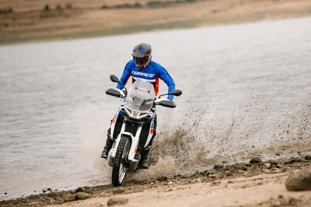 A blue-and-red-clad rider slides a 2022 Aprilia Tuareg 660 on the edge of a sandy lake