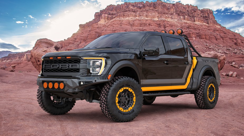 A 2022 Ford F-150 Raptor in a dark grey color with a light grey and yellow racing stripe graphics. The truck is lifted with off road wheels and tires. The wheels have bead locks that are yellow and color matched to the graphics. This truck was on display at SEMA in 2021.