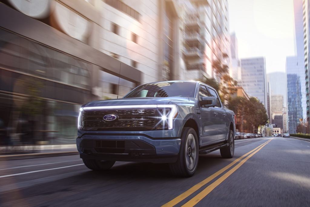 Blue 2022 Ford F-150 Lightning driving down the street, ready to charge battery at EV charging station, Millennials buy more pickup trucks than Boomers, and any other generation.