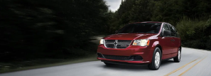 The Dodge Grand Caravan minivan with a red paint color option driving on a highway surrounded by forest trees