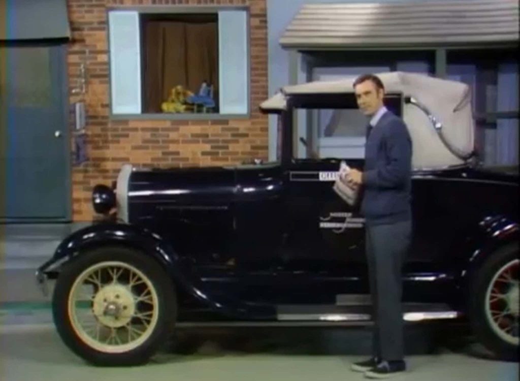 Fred Rogers standing next to a navy blue 1928 Ford Model A Sport Coupe in Mister Rogers' Neighborhood with a house in the background