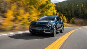 Front view of blue 2023 Kia Sportage Hybrid driving by a forest