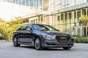 The Genesis G90 luxury full-size sedan with a gray paint color option parked in a building plaza near bushes and trees