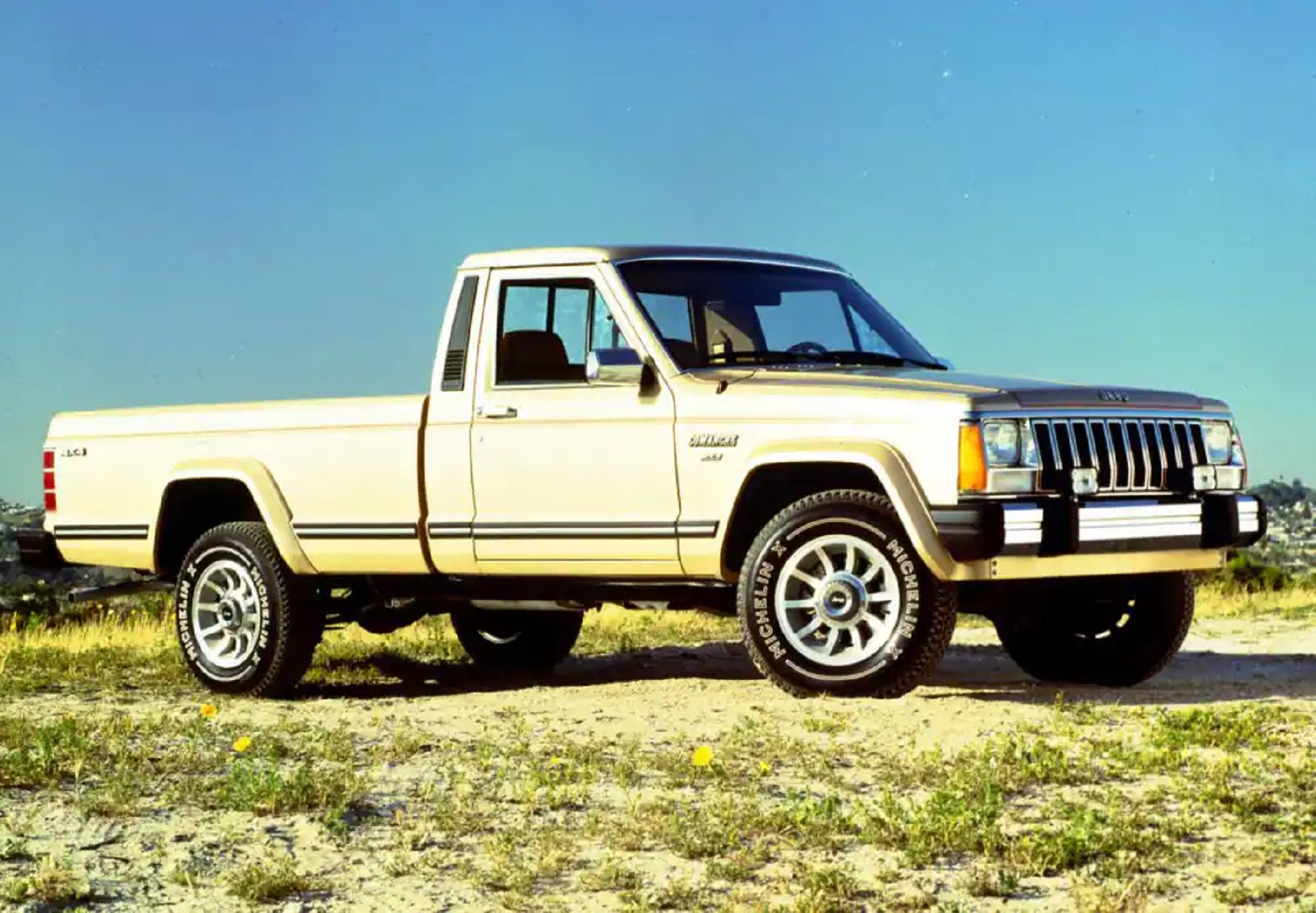 A silver-and-gold Jeep 'MJ' Comanche parked on a sandy grassy hill