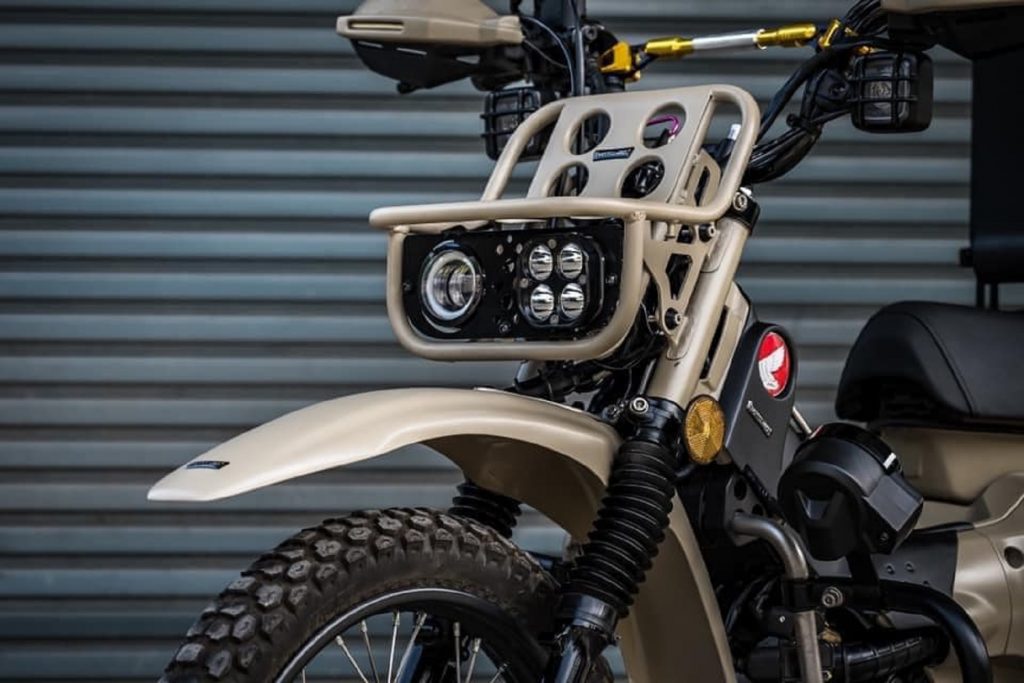 A close-up view of MotolordD's 2021 Honda Trail 125 LED headlight braket and luggage rack on a sand-colored Trail 125