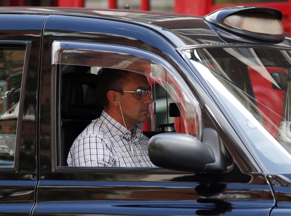 A man driving a taxi in London wears headphones while driving, which could be illegal in some areas.