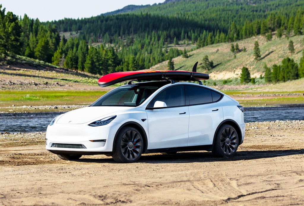 White Tesla Model Y parked at a lake with a red surfboard on top. Tesla has increased the price of the Model Y and it could be because of new EV tax credits